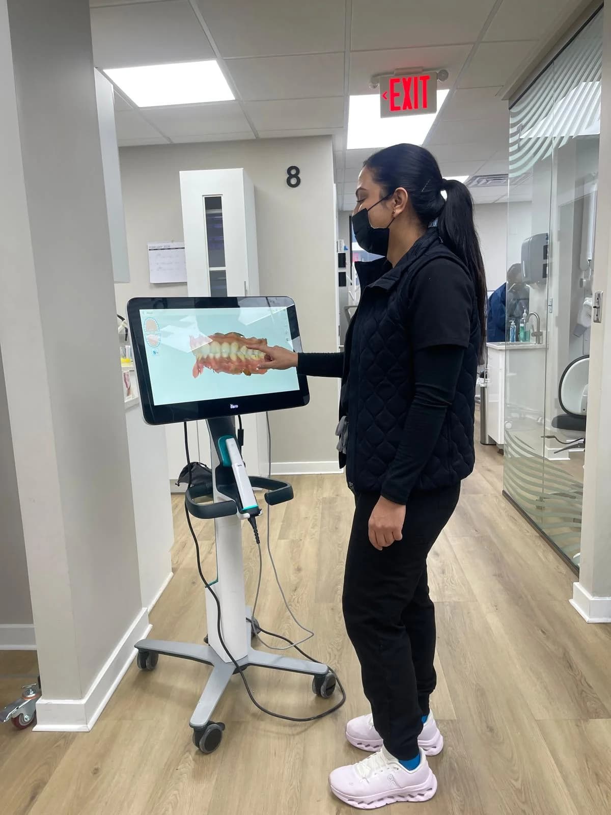 A masked member of the team at the digital scanner cart in the corridor, with a three-dimensional scan of an upper and a lower arch closed together on the screen.